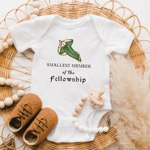 Puede incluir: Body de bebé blanco con el texto "SMALLEST MEMBER of the Fellowship" y un diseño de hoja verde. La imagen incluye zapatos de bebé marrones, un chupete y un clip de chupete con cuentas. Los artículos están colocados sobre una estera de mimbre tejida.