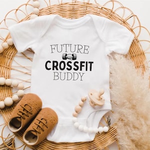 May include: A white baby bodysuit with the text "FUTURE CROSSFIT BUDDY" in black. The image includes brown baby shoes, a wooden dummy, and a beaded necklace. The items are arranged on a woven wicker background.