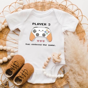 May include: White baby onesie with the text "PLAYER 3 has entered the game" and a cartoon game controller. Includes brown baby shoes and a pacifier with a wooden bead chain. The onesie is on a woven wicker background.