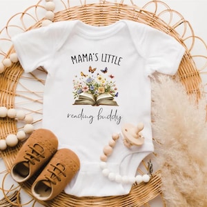 Puede incluir: Body de bebé blanco con el texto "Mama's Little reading buddy" y un diseño floral de libro. La imagen también incluye zapatos de bebé marrones y un chupete con una cadena de cuentas de madera. Los artículos están dispuestos sobre una superficie de mimbre tejido.