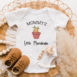 Può includere: Body da bambino bianco con la scritta "Mommy's Little Mandrake" e un'illustrazione di una pianta di mandragora. Include scarpine marroni e un ciuccio con una catena di perline. Gli oggetti sono disposti su una superficie di vimini intrecciato.