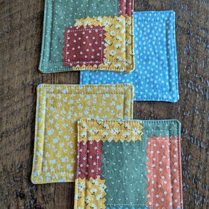 May include: Four square fabric coasters with a patchwork design. The coasters feature a mix of floral, polka dot, and solid patterns in shades of yellow, green, red, and blue. Each coaster is approximately 4 inches square.