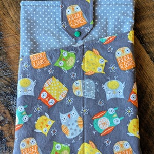 May include: A gray fabric book cover with a flap closure. The cover features a pattern of colorful owls and small white flowers on a gray background. The flap is secured with a green button.