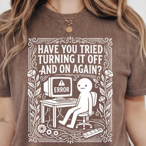 Peut inclure: T-shirt marron avec un motif blanc. Le motif présente le texte "HAVE YOU TRIED TURNING IT OFF AND ON AGAIN ?" au-dessus d'un ordinateur avec un message d'erreur et un bonhomme allumette. Le motif est encadré d'éléments floraux.