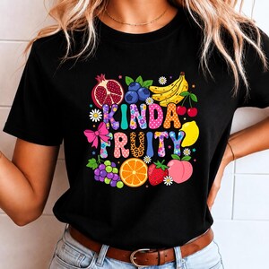 Peut inclure: T-shirt noir avec un motif graphique coloré de fruits, dont une grenade, des myrtilles, des bananes et des cerises. Le texte "KINDA FRUITY" est intégré au design avec différentes polices et motifs.