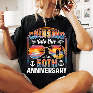 Peut inclure: Un t-shirt noir avec une impression graphique portant le texte "CRUISING Into Our 50TH ANNIVERSARY". Le motif montre des lunettes de soleil reflétant une plage au coucher du soleil avec un yacht et un couple, encadré par des ancres et des rayures rétro.