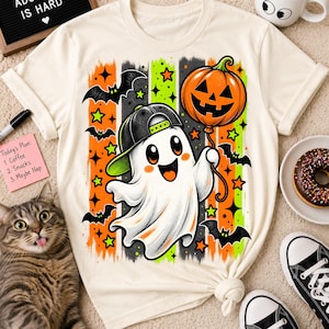 Peut inclure: Un t-shirt couleur crème arborant un motif de fantôme blanc portant une casquette de baseball noire et tenant un ballon citrouille orange. Le design comprend des chauves-souris noires, des traits verticaux orange et verts, et des étoiles.
