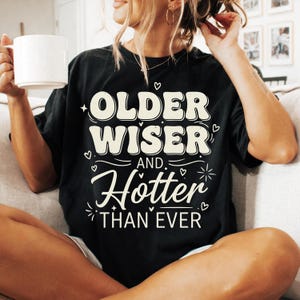 May include: Black t-shirt with the text "OLDER WISER AND Hotter THAN EVER" in a retro font. The design includes small heart and starburst graphics. The shirt is a casual style, suitable for everyday wear.