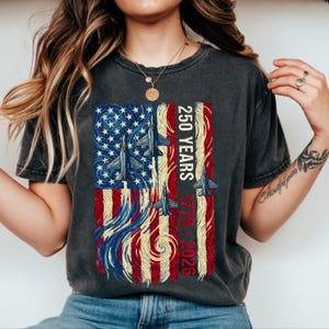 Peut inclure: T-shirt gris foncé avec un motif de drapeau américain stylisé, des avions de chasse et le texte "250 YEARS 1776-2026". Le motif du drapeau utilise les couleurs rouge, blanc et bleu.