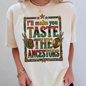 Peut inclure: T-shirt crème avec un motif graphique coloré. Le design affiche le texte "I'll make you TASTE THE ANCESTORS" en rouge, noir, vert et jaune, encadré d'éléments d'art tribal.