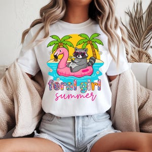Peut inclure: T-shirt blanc avec un motif coloré d'un raton laveur portant des lunettes de soleil, se relaxant sur un flotteur flamant rose dans l'océan. Le texte "feral girl summer" est en dessous. Des palmiers et des fleurs complètent le design.