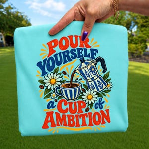 Peut inclure: Un t-shirt bleu clair avec une impression graphique représentant une cafetière italienne versant du café dans une tasse, entourée de marguerites blanches et de grains de café. Le texte indique "POUR YOURSELF A CUP OF AMBITION" en lettres rouges et bleues.