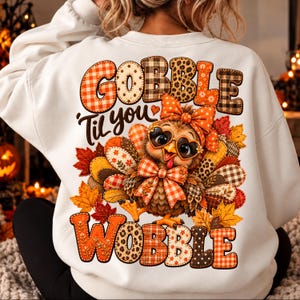 May include: A white crewneck sweatshirt featuring a graphic on the back. The design shows a turkey wearing heart-shaped glasses and a bow, surrounded by autumn leaves. The text reads GOBBLE 'til you WOBBLE in patterned orange and brown letters.