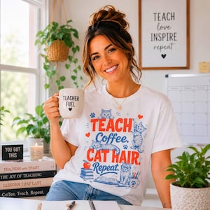 Op de afbeelding: Een wit T-shirt met korte mouwen en een grafische print met de tekst "TEACH Coffee. CAT HAIR. Repeat." omringd door illustraties van katten, boeken, een appel en een pennenhouder. Het T-shirt wordt gecombineerd met een witte mok met "TEACHER FUEL."