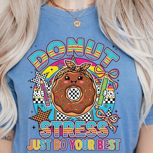 Peut inclure: T-shirt bleu clair avec un motif coloré. Le motif comprend le mot "DONUT" en arc-en-ciel, un personnage de donut souriant et les mots "STRESS JUST DO YOUR BEST". Le donut porte un bandeau léopard.