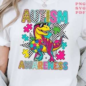 May include: White t-shirt with a colorful graphic design promoting autism awareness. The design features the word "AUTISM" in a bold, multi-colored font, and a cartoon dinosaur wearing sunglasses and holding a puzzle piece. The word "AWARENESS" is also displayed.
