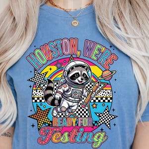 Könnte beinhalten: Hellblaues T-Shirt mit einem farbenfrohen Aufdruck. Der Aufdruck zeigt einen Waschbären in einem Astronautenanzug, der einen Bleistift hält, mit dem Text "Houston, We're Ready For Testing" in einem Regenbogen-Design. Das Design enthält Sterne und einen Planeten.