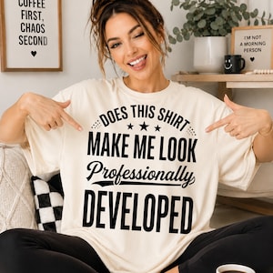 Peut inclure: Un t-shirt à manches courtes de couleur crème avec un texte noir indiquant : DOES THIS SHIRT MAKE ME LOOK Professionally DEVELOPED. Le motif comprend trois petites étoiles centrées entre les deux premières lignes de lettres grasses.