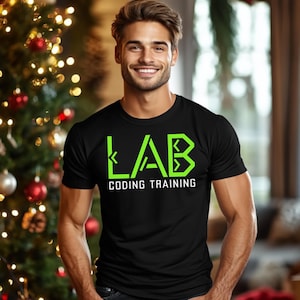 May include: Black t-shirt with the word "LAB" in bright green, with the words "CODING TRAINING" below. The person is smiling in front of a Christmas tree with red and gold ornaments.