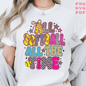 Peut inclure: T-shirt blanc avec un graphisme coloré qui dit "ALL SOFTBALL ALL THE TIME". Le motif comprend un ballon de softball, des étoiles et les mots dans divers motifs et couleurs, dont le jaune, le rose et le bleu. Le t-shirt est un vêtement décontracté.