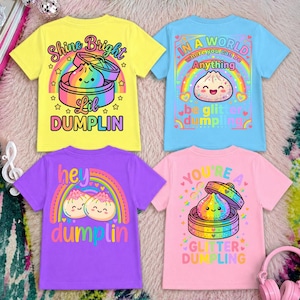Könnte beinhalten: Vier farbenfrohe Kinder-T-Shirts. Die Shirts sind gelb, blau, lila und rosa. Jedes Shirt zeigt eine Knödel-Grafik mit Text. Das gelbe Shirt sagt "Shine Bright Lil Dumplin". Das blaue Shirt sagt "In a world where you can be anything be glitter dumplin".