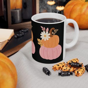 May include: White ceramic mug with a black background featuring a stacked pumpkin design in shades of orange and pink, with floral accents. The mug is filled with a dark liquid and is surrounded by Halloween-themed candies.