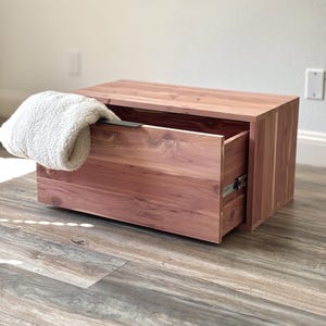 May include: A rectangular cedar wood storage box with a single pull-out drawer featuring a black metal handle. A folded white textured fleece blanket rests on the top left corner of the drawer front, partially covering the black metal handle.