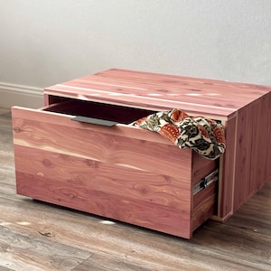 May include: A rectangular cedar wood storage chest with a natural reddish-brown grain pattern. The chest features a single pull-out drawer with a thin black metal handle. A patterned fabric with orange and green floral motifs rests inside the drawer.