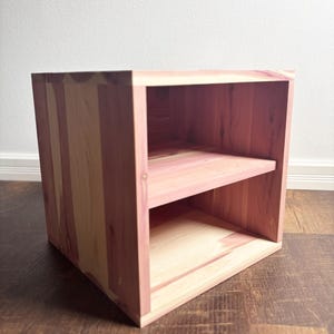 May include: A wooden cube-shaped shelf unit with two shelves. The unit is constructed from light-colored wood with reddish-pink tones. The shelves are evenly spaced within the cube, creating three open compartments. The wood grain is visible.