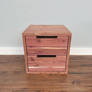 May include: A two-drawer wooden nightstand or small cabinet. The cabinet is made of reddish-brown wood with visible grain patterns. It has two drawers with black, horizontal handles. The cabinet is square and sits on a wooden floor.
