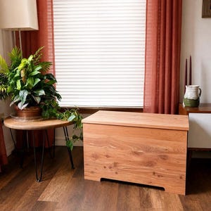 May include: A wooden storage chest with a natural finish sits on a hardwood floor. The chest has a rectangular shape with a flat top and a recessed base. The room features a round side table with a plant, a lamp, and a window with blinds.