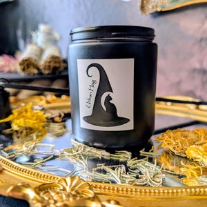 May include: A black jar with a white label featuring a witch hat and cat silhouette, with the text "Urban Hag." The jar sits on a gold-rimmed tray with dried flowers and herbs. A small black bell is also visible.