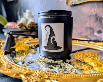 May include: A black jar with a white label showing a witch's hat and cat silhouette, with the text "Urban Hag." The jar is on a gold-edged tray with dried flowers and herbs. A small black bell is also visible.