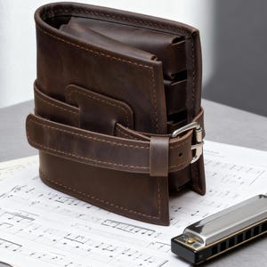 Puede incluir: Una cartera de cuero marrón con cierre de hebilla, sobre partituras. Una armónica plateada y negra está cerca. La cartera mide aproximadamente 13 cm de alto y 10 cm de ancho.