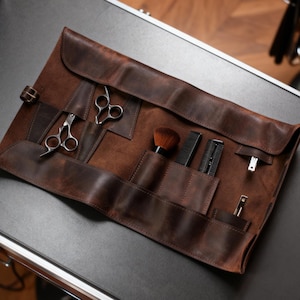 Puede incluir: Un estuche de herramientas de barbero de cuero marrón, abierto sobre una superficie negra. Contiene tijeras, peines y un cepillo. El cuero tiene un aspecto envejecido. En el fondo, una silla de barbero y un suelo de madera. Diseñado para herramientas de barbero.