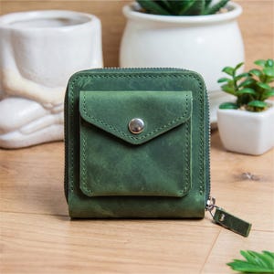 May include: A square, forest green leather wallet with a small front pocket secured by a silver snap. The wallet has a wraparound zipper and a matching green leather pull tab. The wallet is sitting on a wooden surface.
