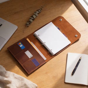 May include: A brown leather binder with a three-ring mechanism, open to reveal blank white paper. The binder has pockets for cards and papers. A pen and notebook are on the wooden surface. A laptop is visible in the background.