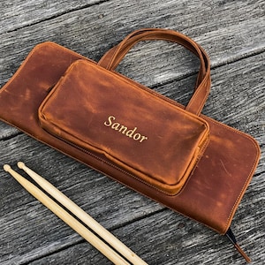Op de afbeelding: Een bruine leren drumstokkenkoker met een bijpassende handgreep en een kleiner zakje. Het woord "Sandor" is in goud op het zakje geëmbosseerd. Twee houten drumstokken liggen naast de koker op een houten oppervlak.