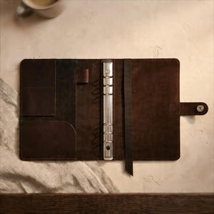May include: A brown leather binder, open and laid flat, featuring a silver ring mechanism. The binder has pockets and a pen holder. A dark brown strap and a snap closure are visible. The binder is on a light-coloured surface.