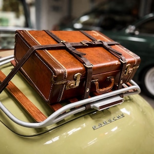 以下が含まれることがあります： ダークブラウンのストラップとバックルで固定された、ヴィンテージの茶色の革製スーツケースが、シルバーの荷物ラックに設置されています。 スーツケースにはハンドルがあり、明るい緑色の車の上にあります。