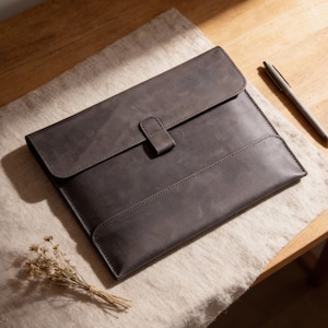 May include: A gray leather laptop sleeve with a flap closure and a tab. The sleeve is rectangular and has a stitched border. A silver pen rests on the wooden surface next to the sleeve. A small bundle of dried flowers is also present.