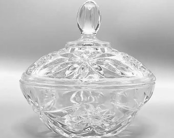 May include: A clear, decorative glass candy dish with a lid. The lid features a teardrop-shaped handle and a starburst pattern. The bowl has a rounded shape with a similar starburst design. The dish is made of cut glass.