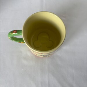 May include: A yellow ceramic mug with a green handle decorated with red and orange floral patterns. The interior of the mug features a Mickey Mouse silhouette at the bottom. The mug has a floral pattern around the rim.