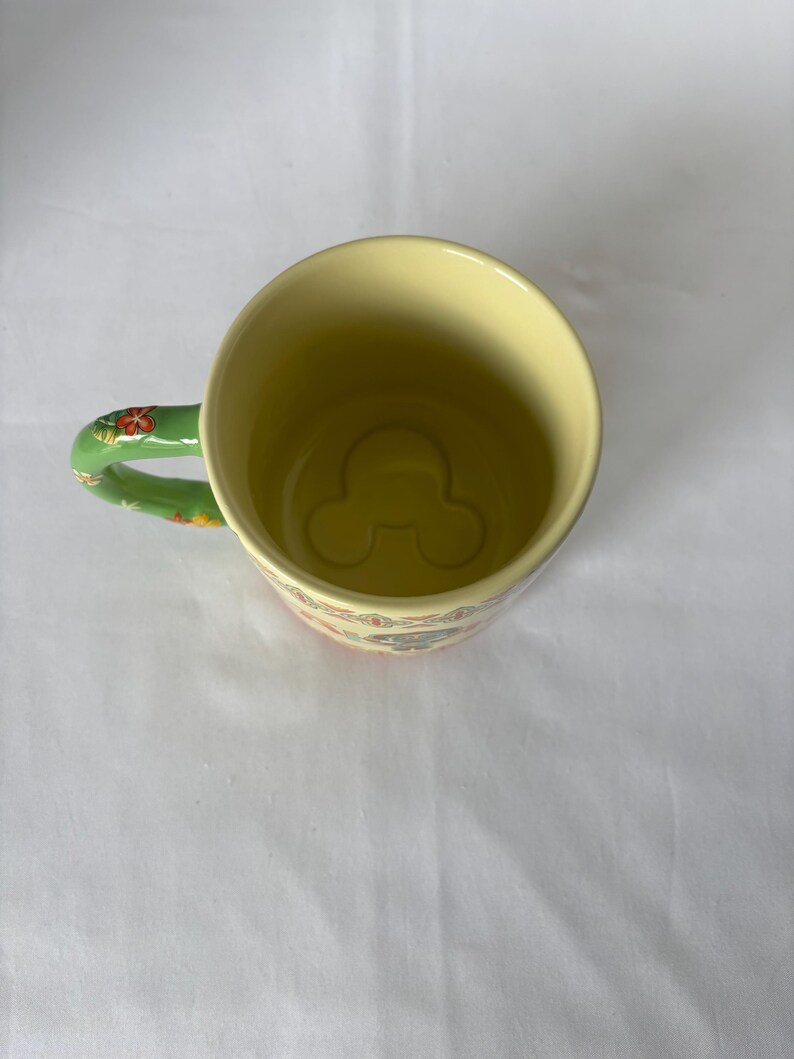 May include: A yellow ceramic mug with a green handle decorated with red and orange floral patterns. The interior of the mug features a Mickey Mouse silhouette at the bottom. The mug has a floral pattern around the rim.