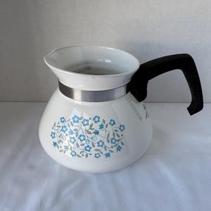 May include: White ceramic coffee pot with a black handle and a silver band. The pot is decorated with a blue floral pattern. The pot has a wide opening and a curved body.