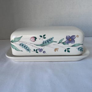 May include: A white ceramic butter dish with a lid, featuring a floral design of purple flowers, green leaves, and pink buds. The dish sits on a matching rectangular base, perfect for serving butter. The design is a classic, elegant style.