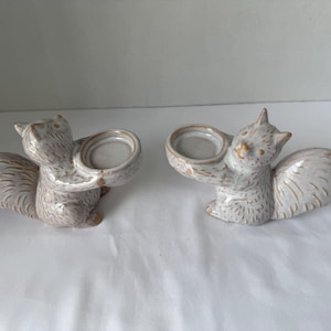 May include: Two ceramic squirrel figurines, each featuring a light beige glaze with darker brown textured detailing. Each squirrel is depicted in a seated position, holding a small, circular, bowl-shaped candle holder in its front paws.