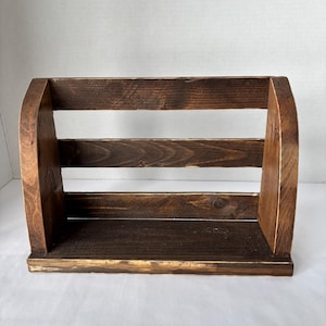 May include: A wooden shelf with a dark brown stain. The shelf has a rectangular base with two vertical side supports and three horizontal slats. The top of the side supports are curved. The wood grain is visible.