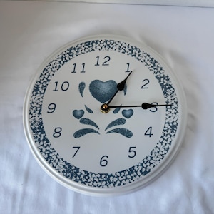 May include: A white wall clock with a blue floral design. The clock face features black hands, a gold center, and blue heart and floral accents. The numbers are black and the border is a dotted blue pattern.