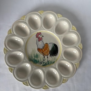 May include: A ceramic egg serving platter with a scalloped edge and a central rooster illustration. The platter has 12 egg-shaped indentations and a yellow border. The rooster is depicted with orange, black, and white feathers.